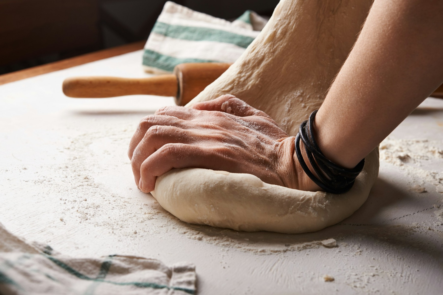 Hands kneading a round of dough on a floured counter, a rolling pin and a folded cloth beside them.